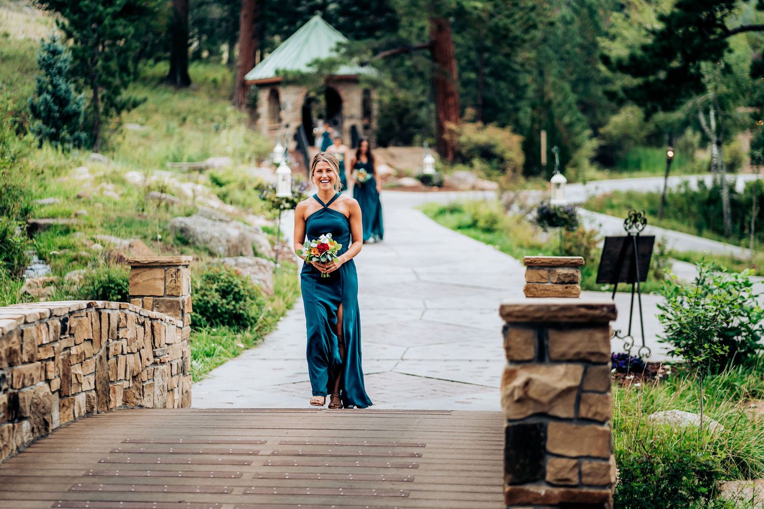 Wedding attendant in a navy blue dress walks down a stone-lined pathway in an outdoor mountain setting.