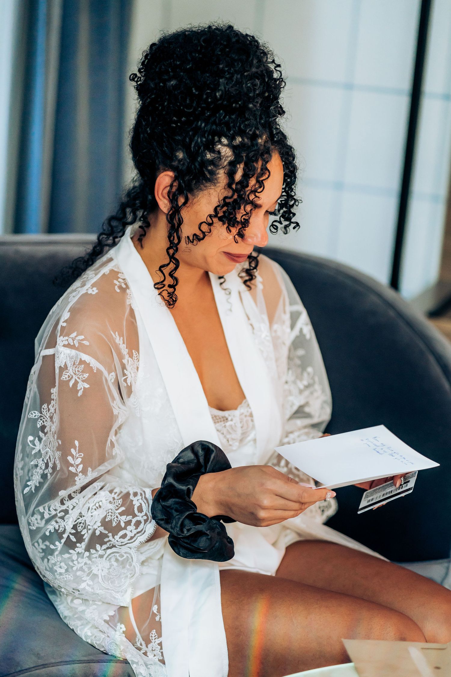 Bride in white lace robe reading while seated on a leather couch.
