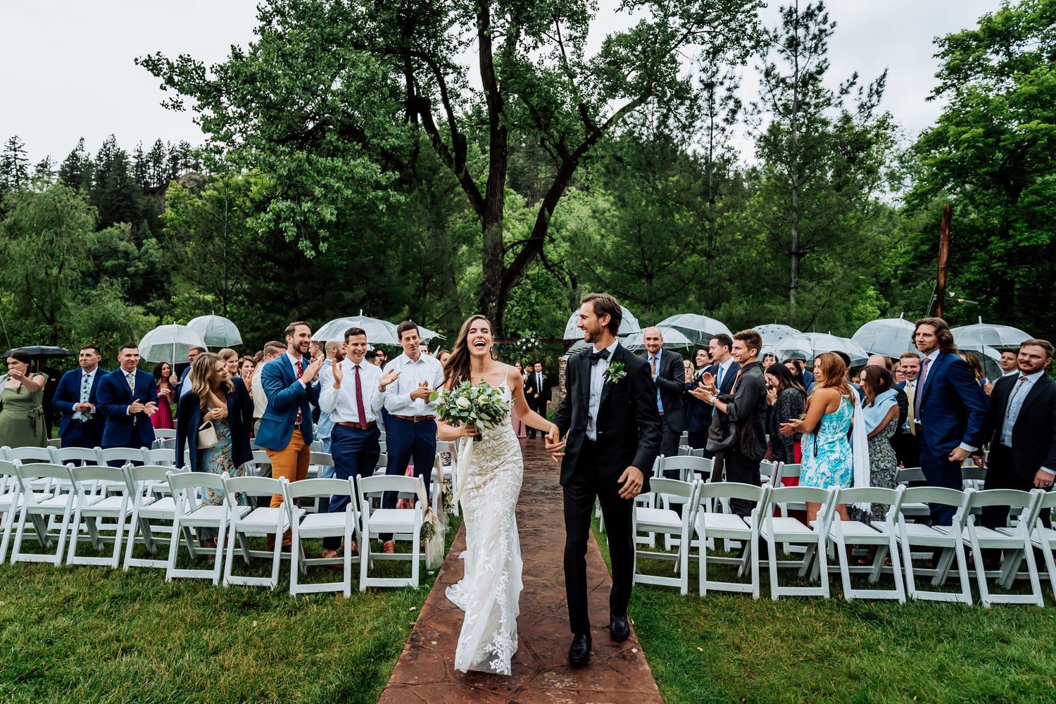 Outdoor wedding ceremony with guests seated in white chairs and umbrellas on a rainy day.