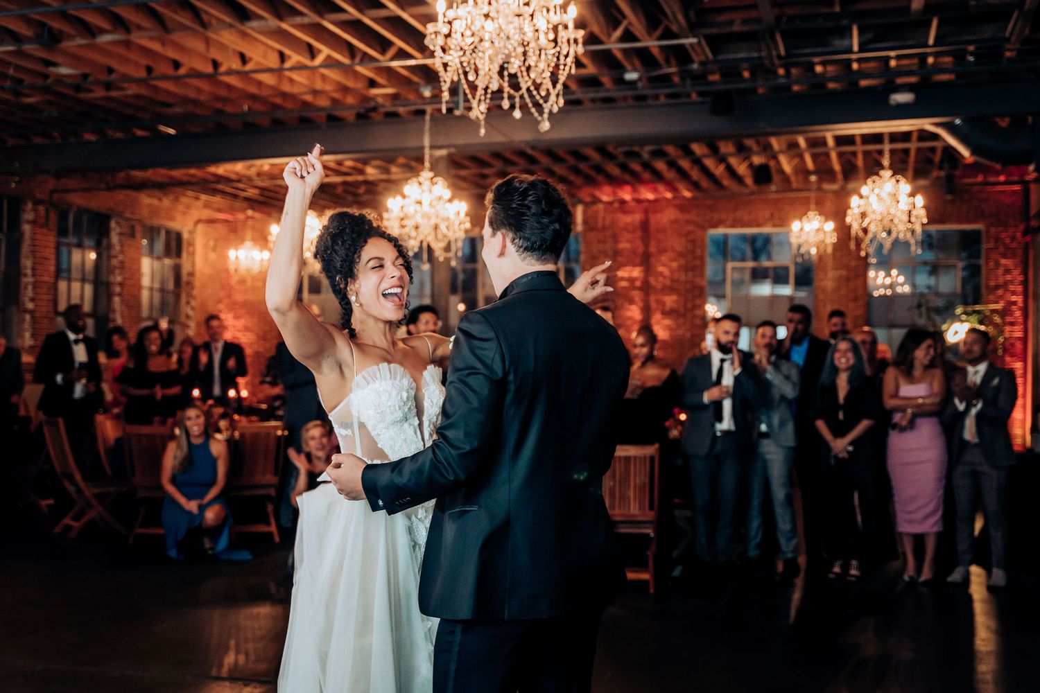 Newlyweds dancing under crystal chandelier in rustic brick venue while guests watch.