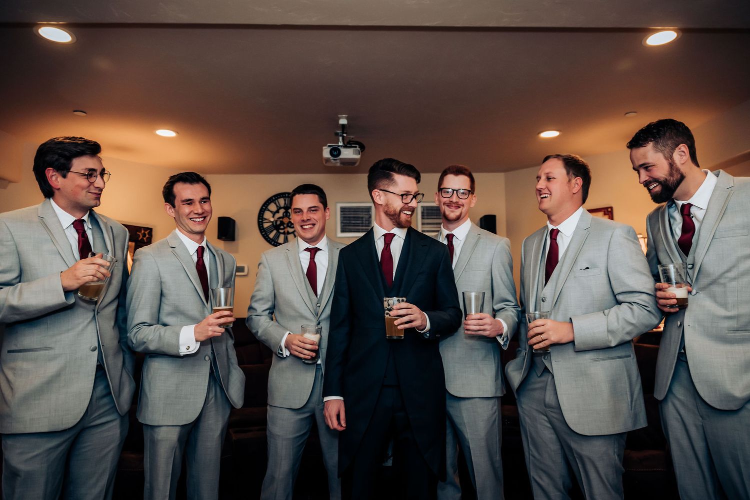 Groomsmen in matching light gray suits sharing drinks and laughter before the wedding ceremony.