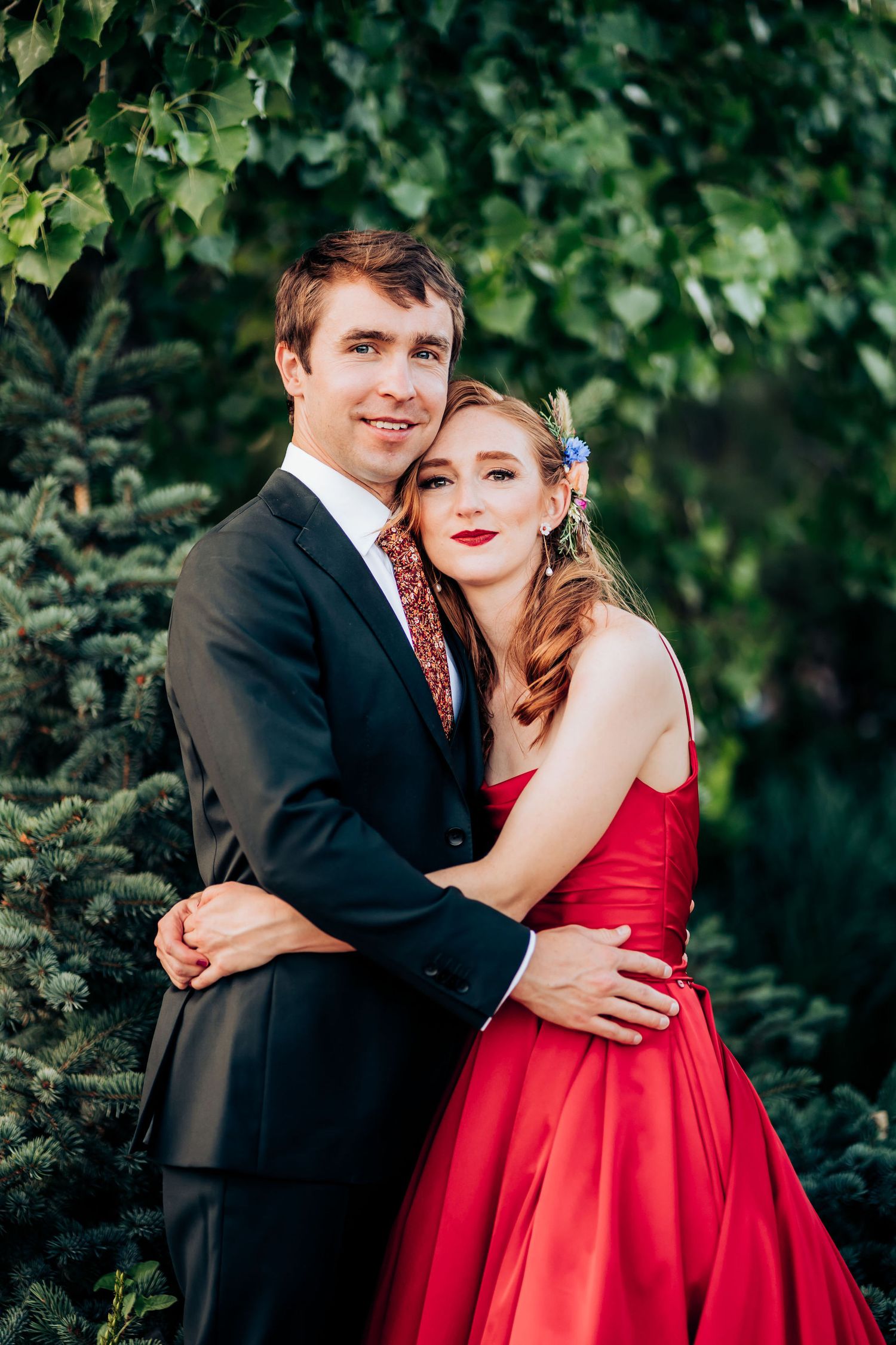 A couple embraces outdoors against green foliage, with her in red gown and him in dark suit.