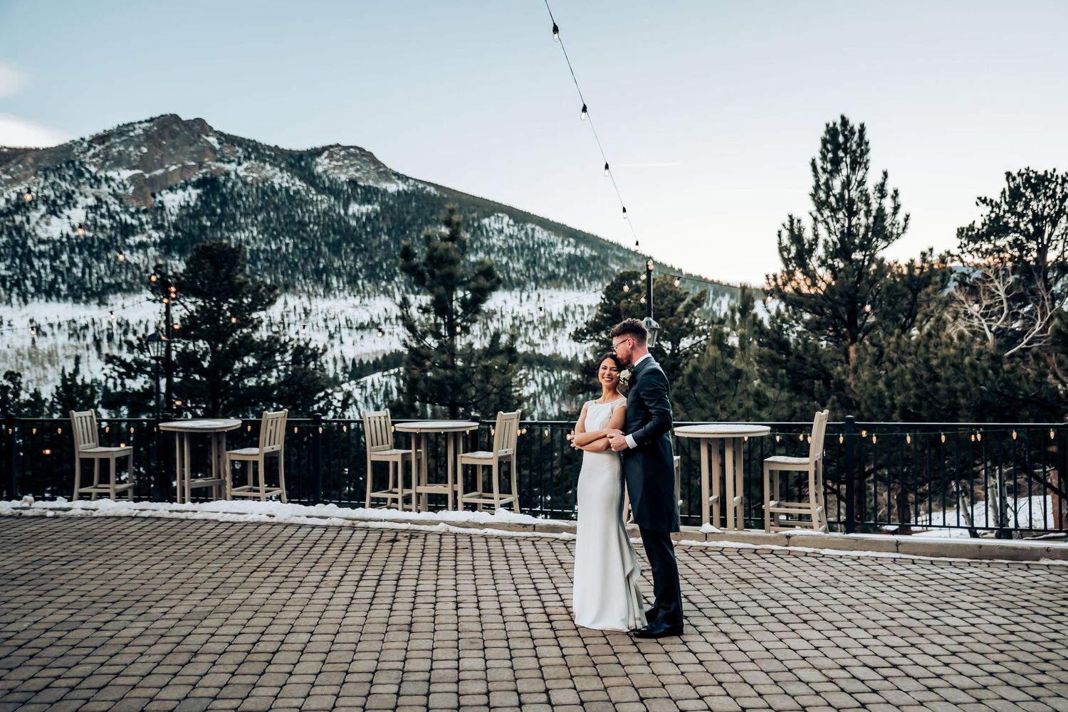 Newlyweds embrace on a terrace overlooking snow-covered mountain peaks at dusk.