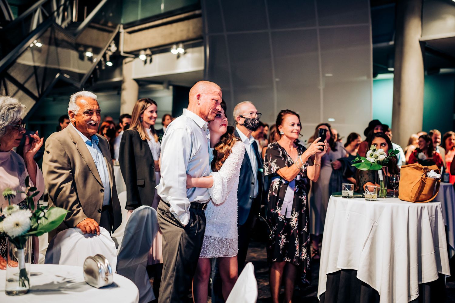 Wedding guests gather around tables during an indoor reception celebration.