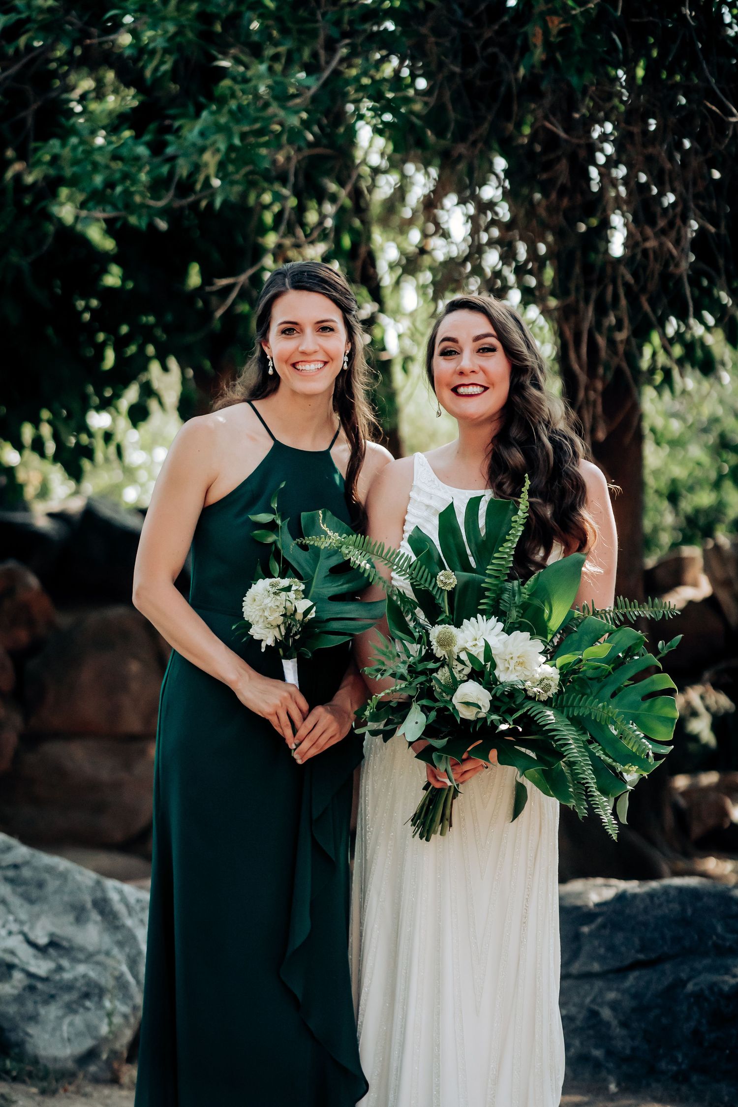 A bridesmaid in emerald green and bride in white hold tropical leaf bouquets against a lush garden backdrop.