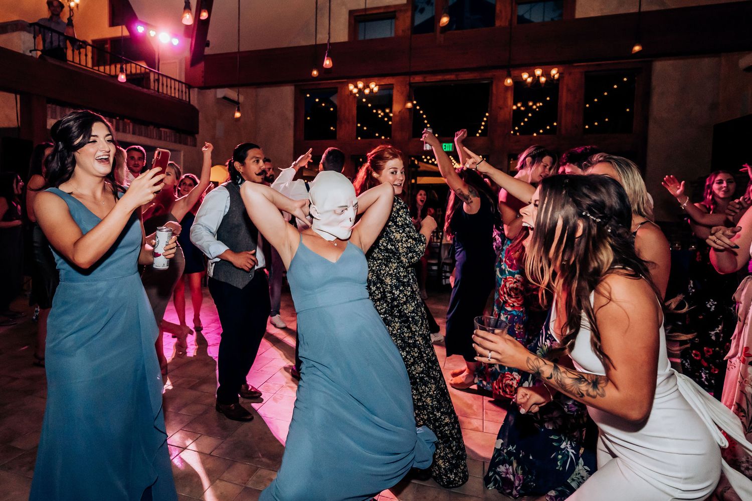 Wedding guests in blue dresses dancing enthusiastically at a reception party with pink mood lighting.