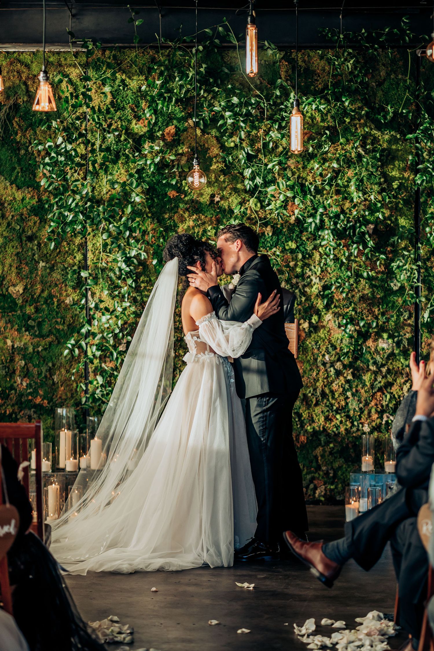 Romantic first kiss moment under hanging lights and ivy wall during wedding ceremony.