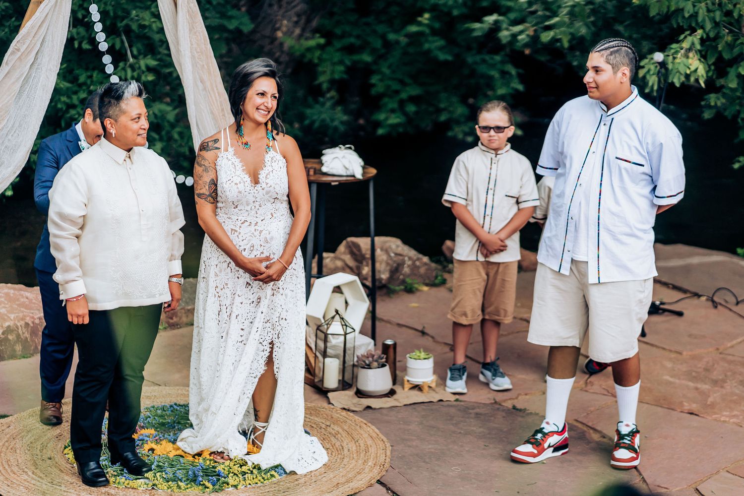 Wedding ceremony participants gather under white drapes in an outdoor garden setting.