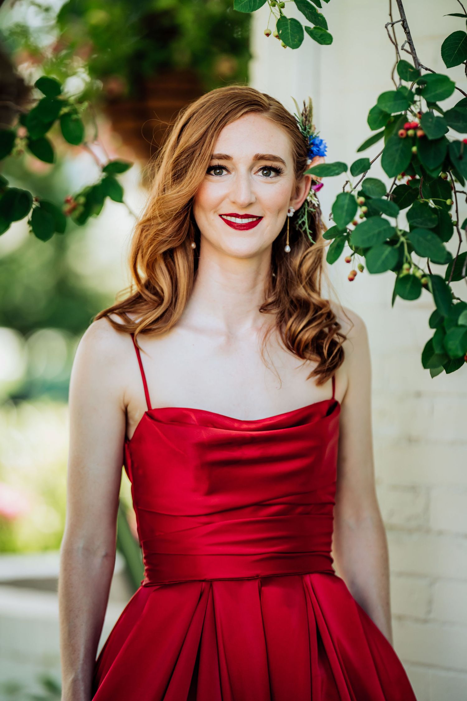 Elegant red formal dress portrait with natural greenery and white wall backdrop.