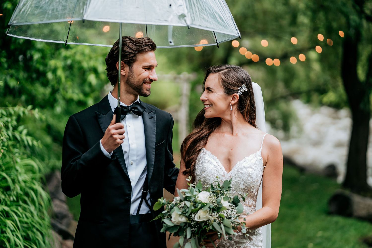 Joyful wedding couple laughing together under a clear umbrella with string lights and greenery in background.