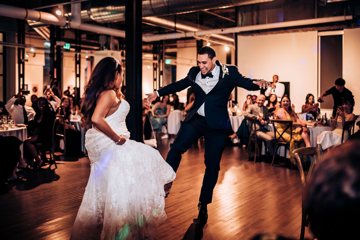 A couple performs an energetic dance move during their wedding reception as guests watch from their tables.