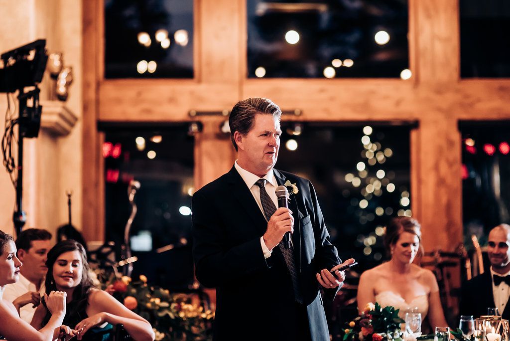 Speaker gives toast at wedding reception with festive lighting and decor.