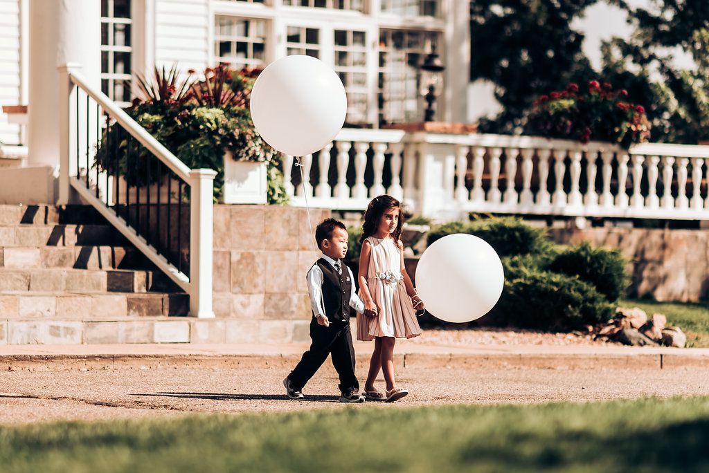 Children carrying white balloons walk across lawn in formal wedding attire.