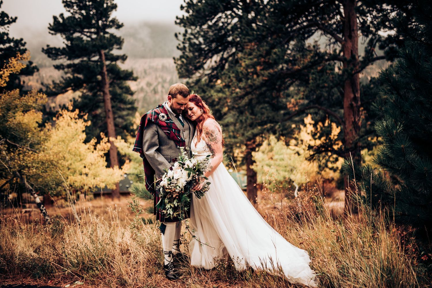 Wedding couple embracing in golden meadow with pine trees and misty mountain backdrop.