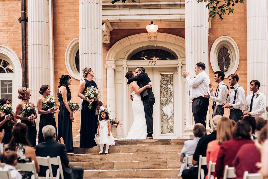 Wedding kiss moment on the steps of a classic brick building with guests watching.