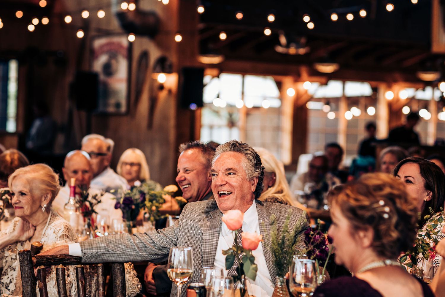 Reception guests celebrating with laughter at tables under warm string lights.