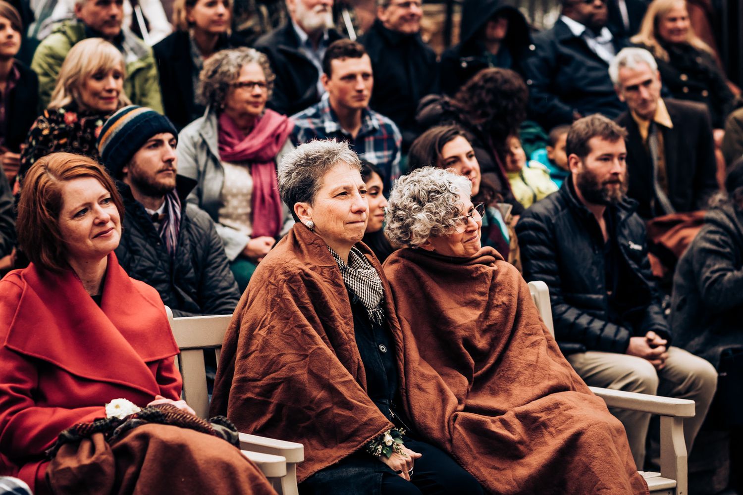 Wedding guests bundled in warm shawls and coats sit in outdoor ceremony chairs.