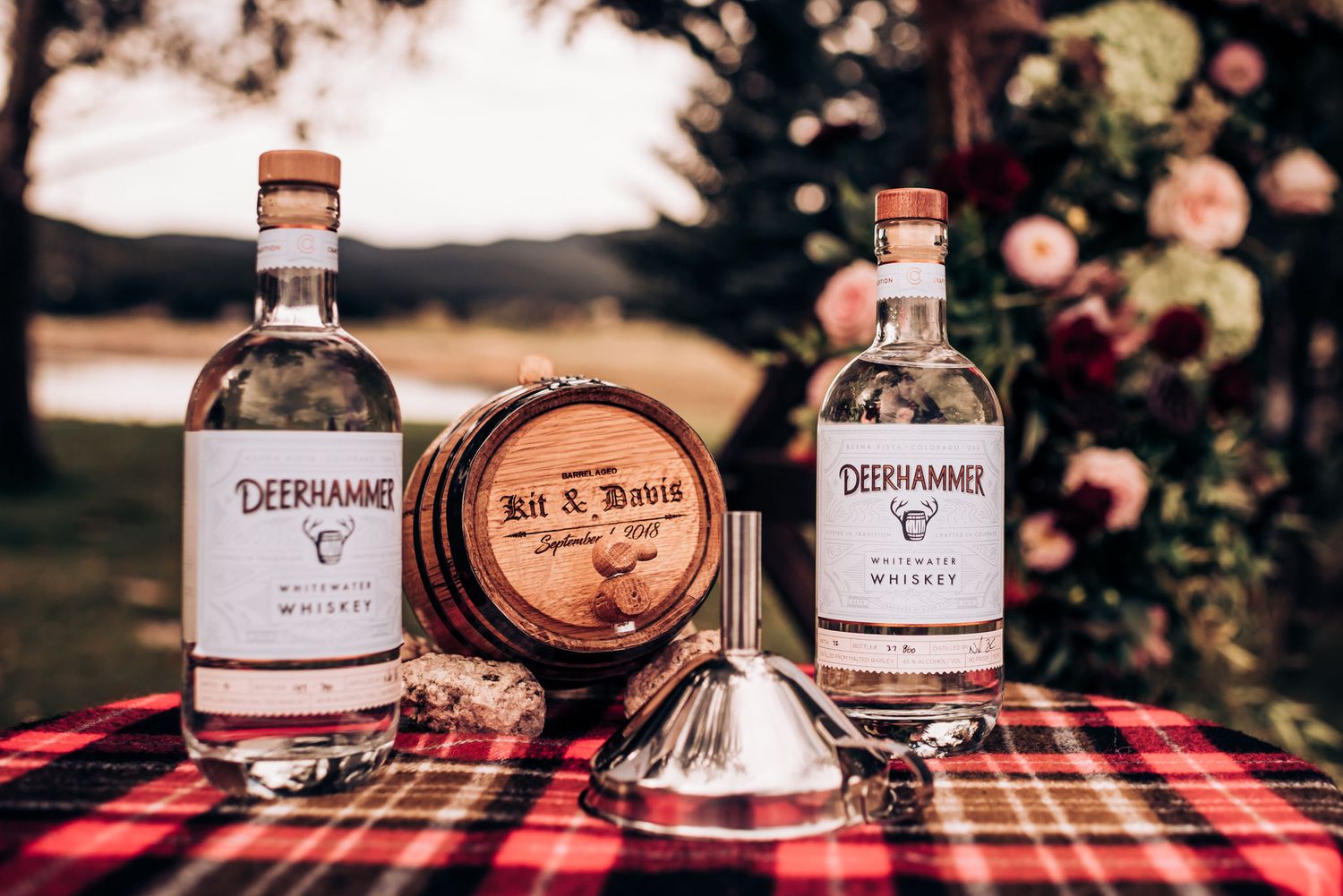 Rustic wedding display with whiskey bottles and small barrel on red plaid tablecloth.