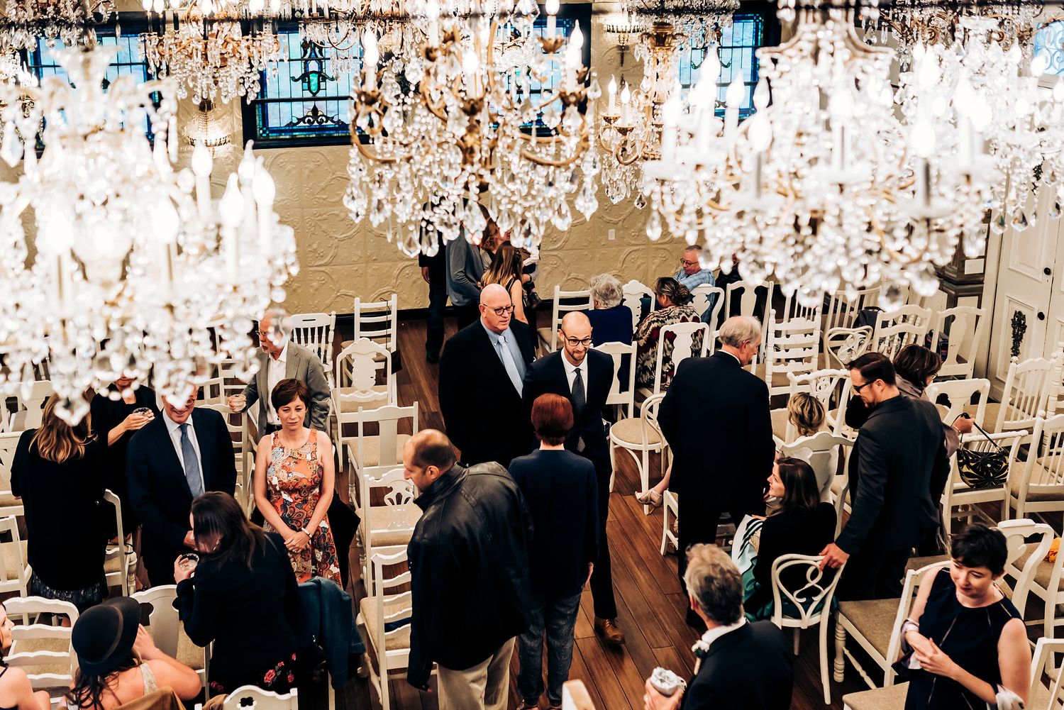 Indoor ceremony space decorated with multiple crystal chandeliers and white chairs.