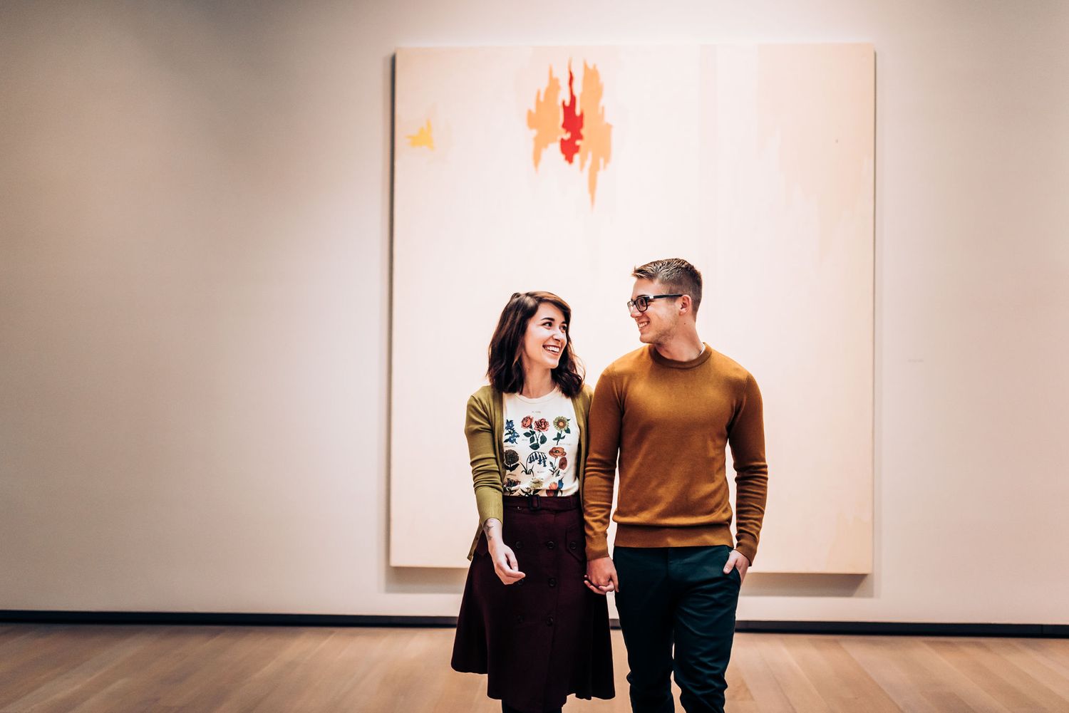 A couple stands in an art gallery viewing a minimalist white and orange painting.