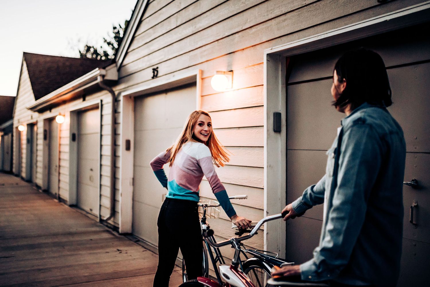 A couple with bikes interacts playfully by garage doors at sunset.