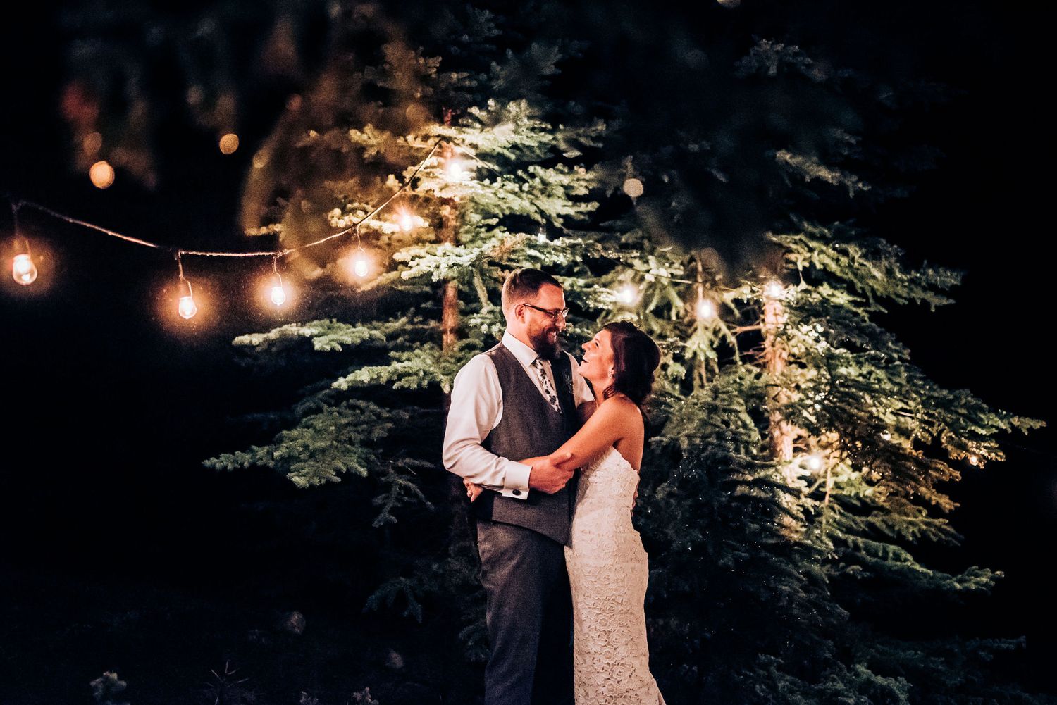 Romantic evening portrait of couple dancing under string lights and trees.