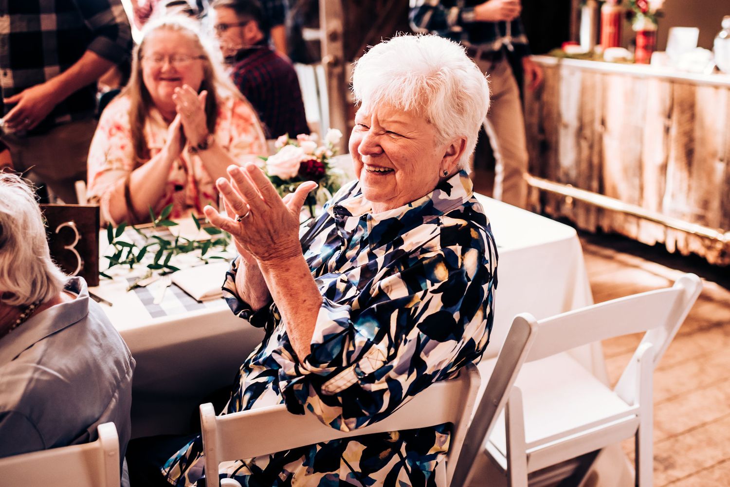 A delighted guest clapping and laughing at a wedding reception in a rustic venue.