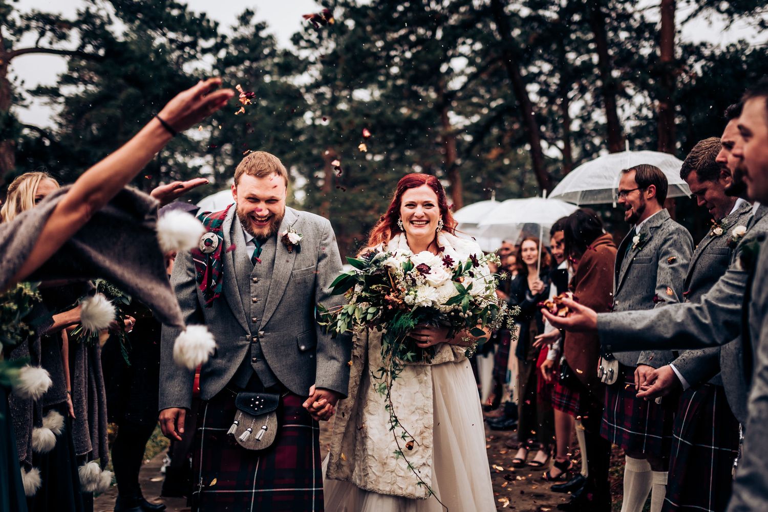Wedding guests throw confetti as newlyweds walk through, with Scottish kilts visible in the crowd.