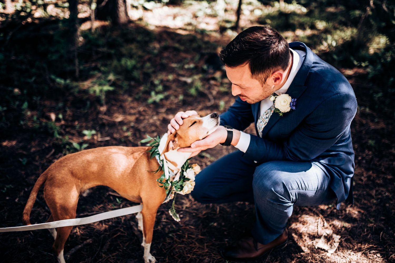 A brown dog wearing a floral collar gets affectionate pets during a forest wedding.
