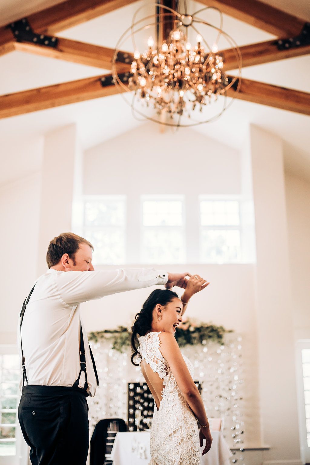 A touching dance moment under a grand chandelier in an elegant white venue with exposed beams.