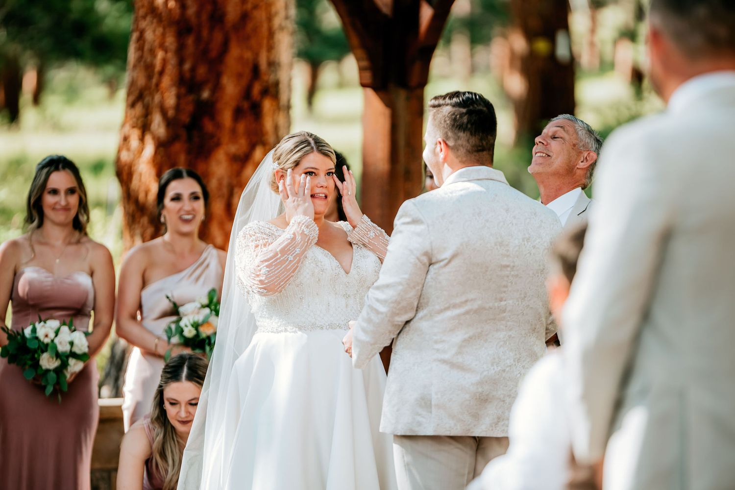 Outdoor wedding ceremony under tall pine trees with attendants in soft pink dresses.