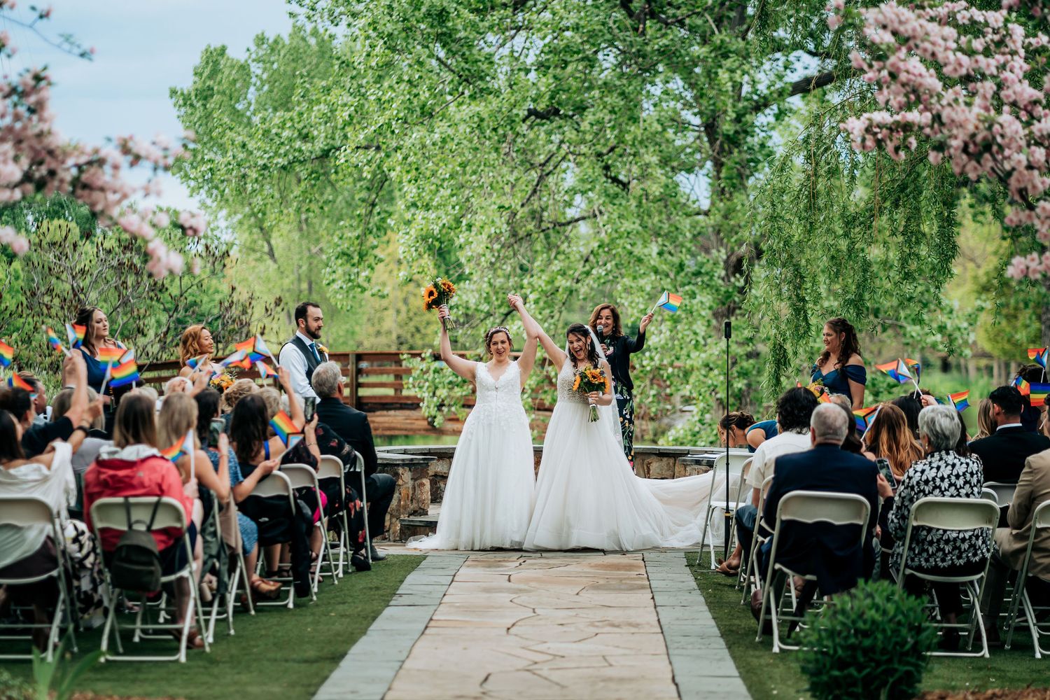 Wedding celebration with guests seated outdoors as couple walks down stone aisle under green trees.
