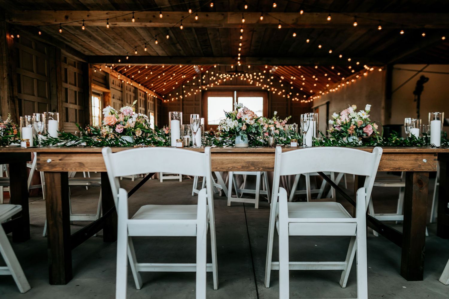Wedding reception setup with white chairs and string lights in rustic wooden venue.