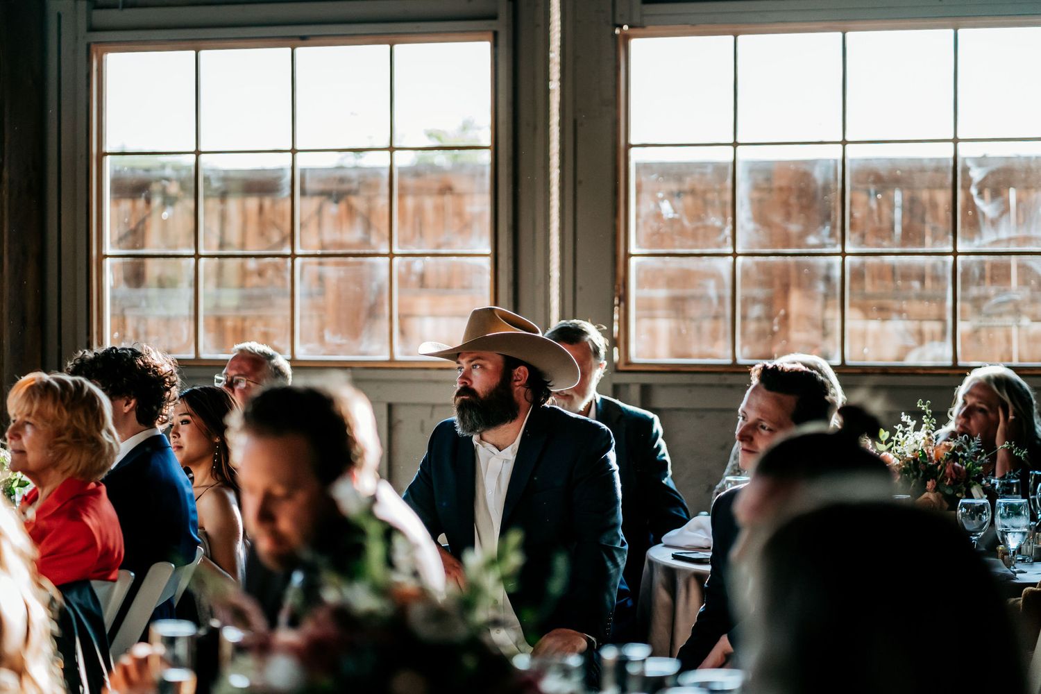 Wedding guests enjoy the festivities at a rustic reception venue with large windows and natural lighting.