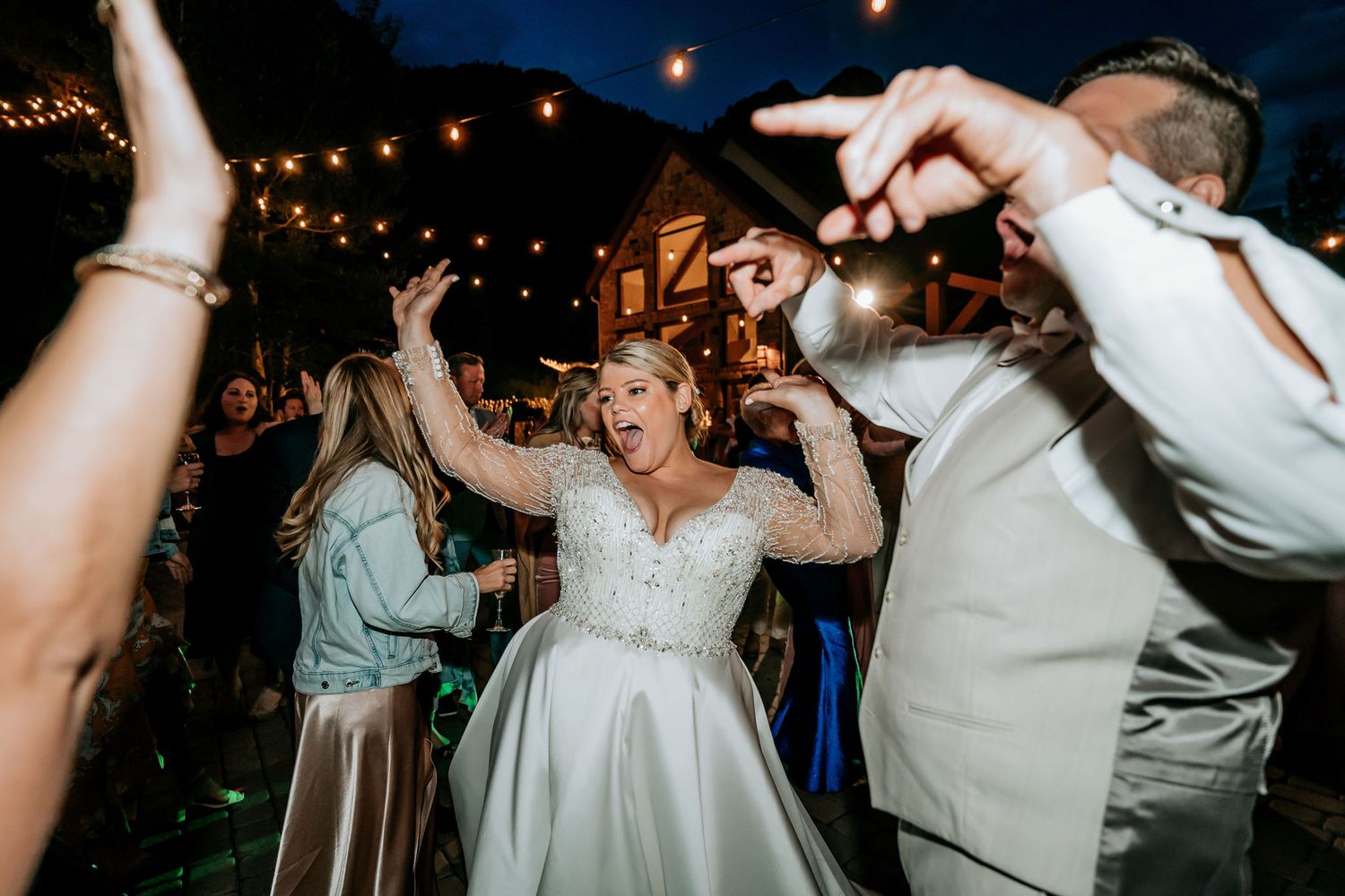 Wedding guests dance joyfully under outdoor string lights at a nighttime reception celebration.