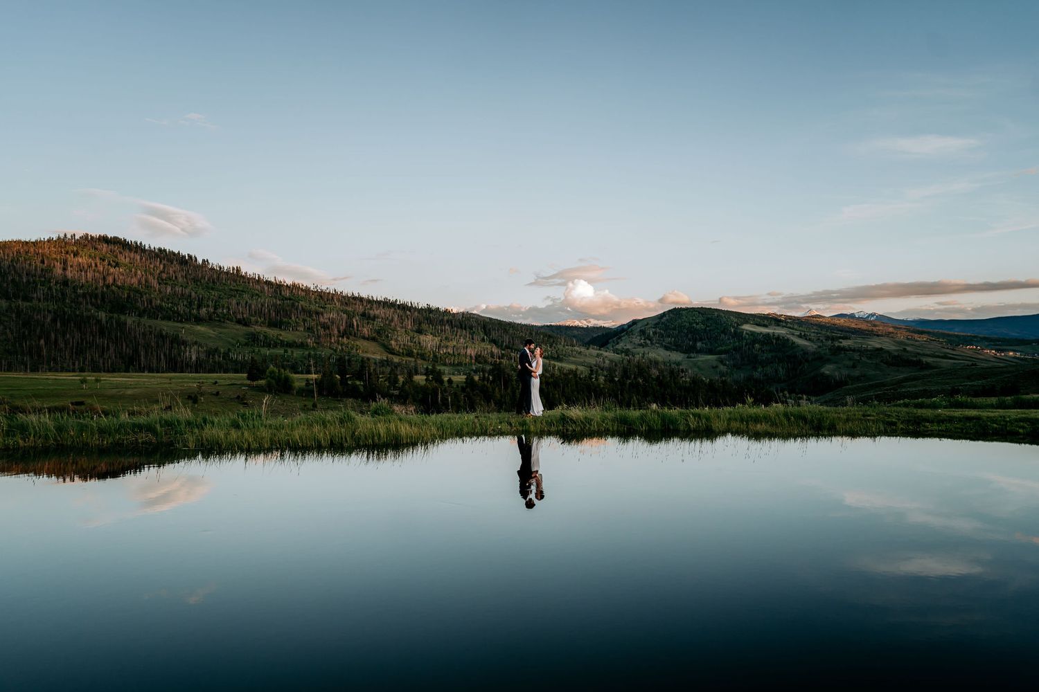 Distant wedding couple silhouetted against mountain landscape with lake reflection.