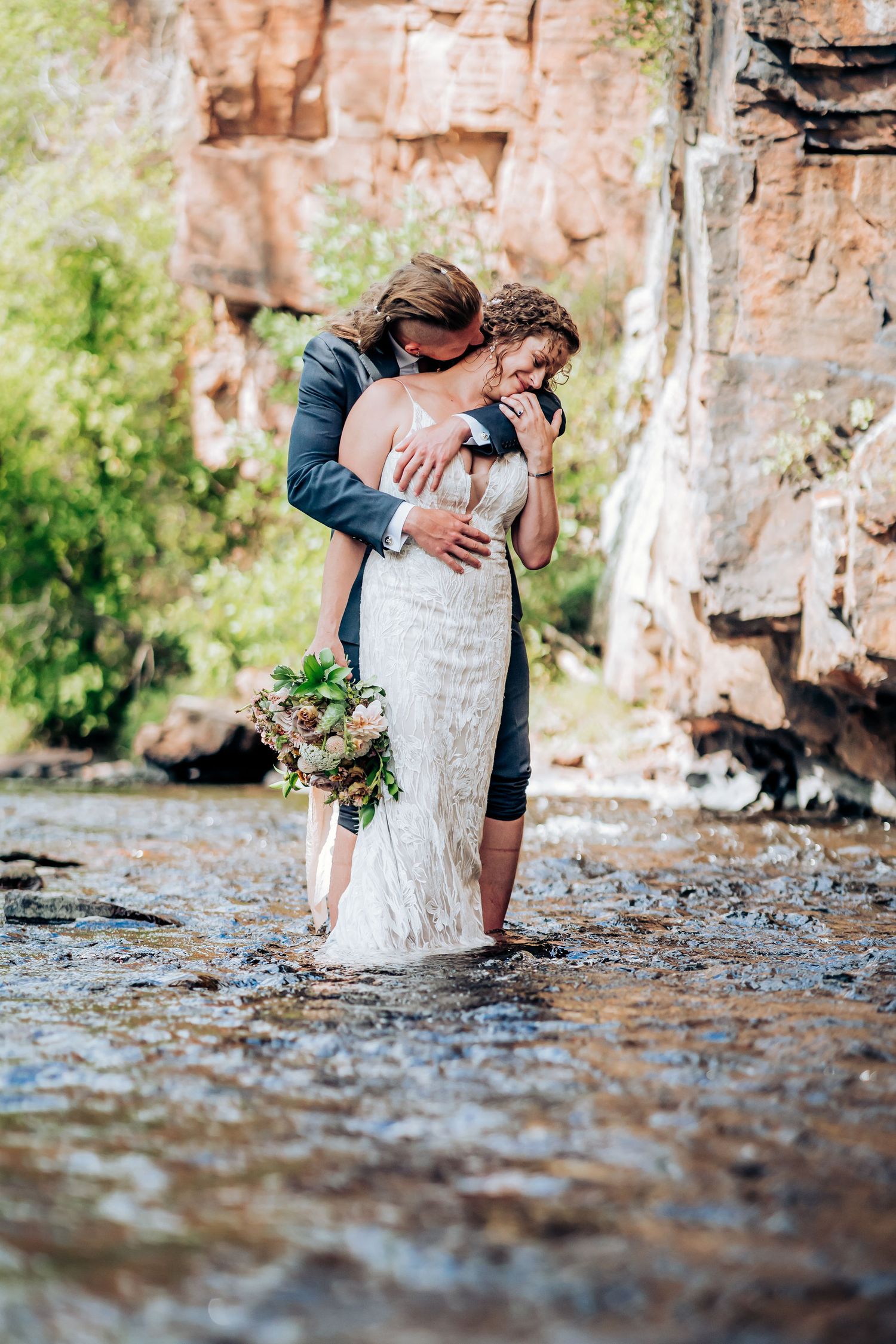 Intimate wedding moment captured in river water with towering cliffs in background.
