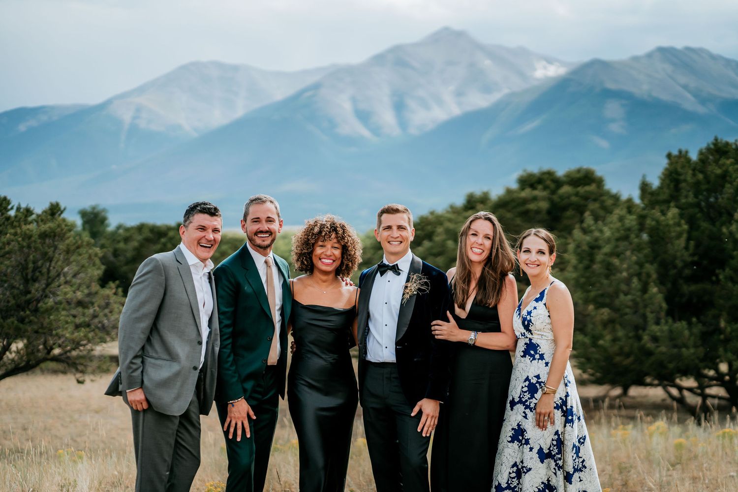 Group of six people in formal attire posing together with mountain landscape behind them.