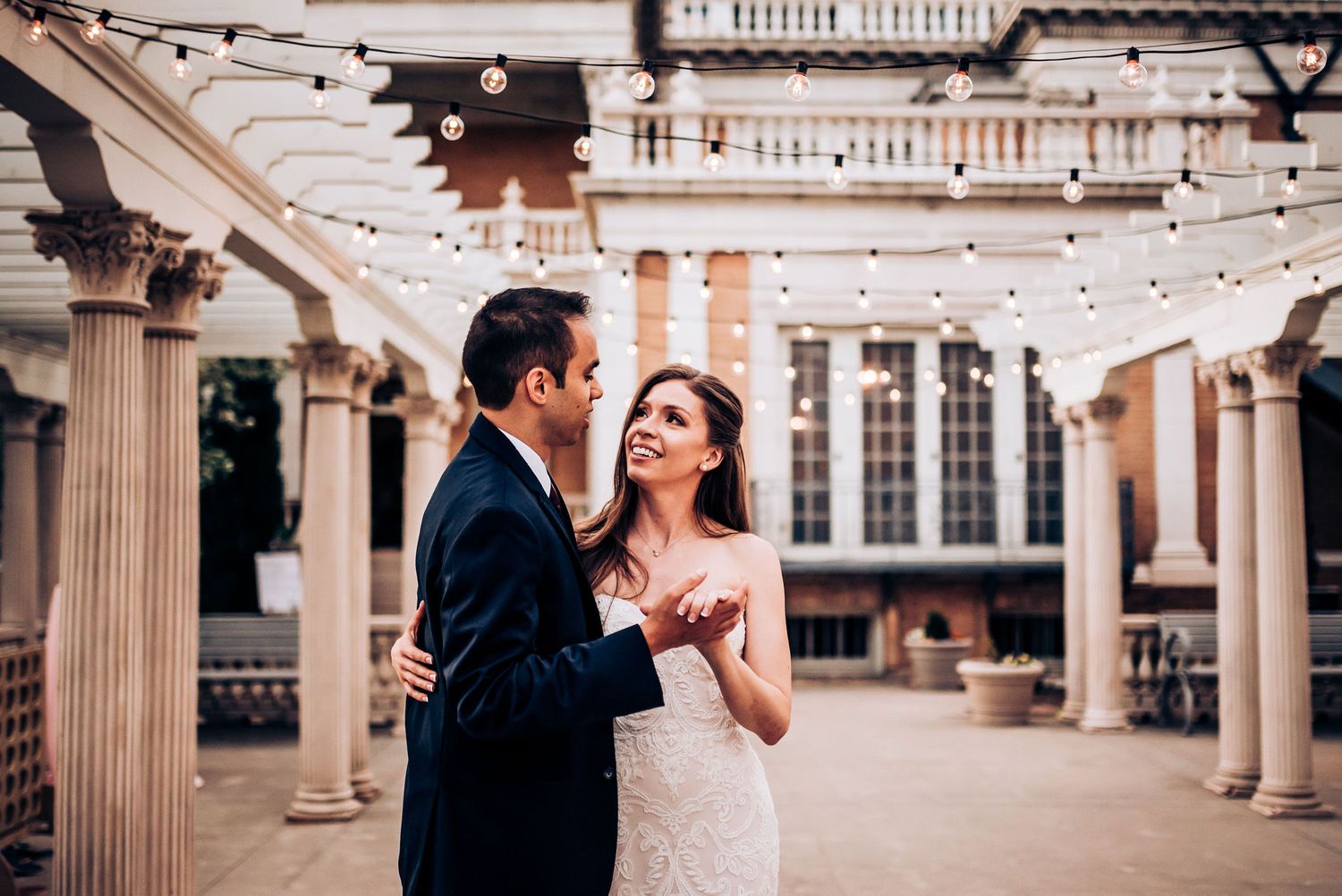 A couple shares an intimate dance moment under string lights and columns in an elegant outdoor venue.
