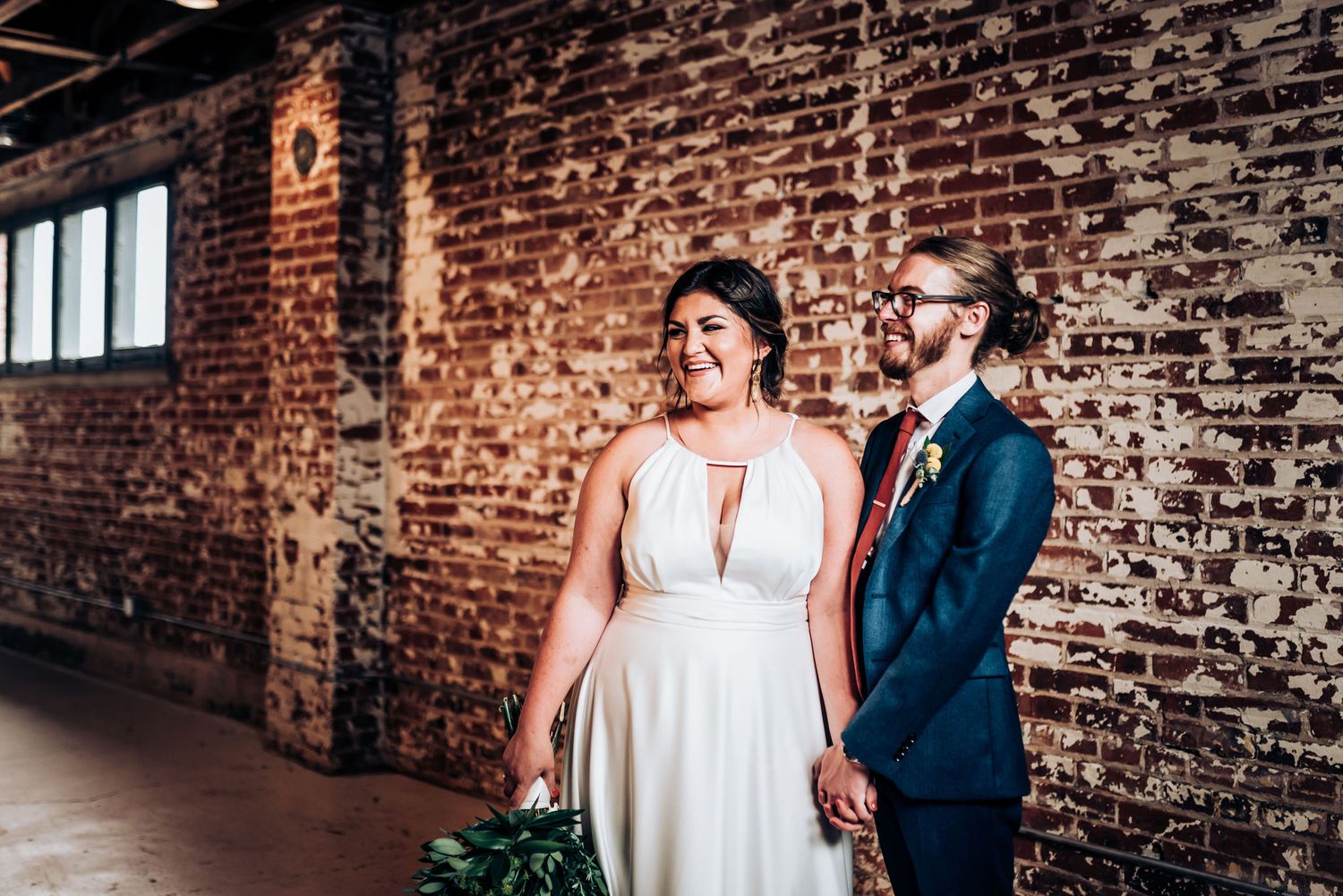 A wedding couple poses against a rustic exposed brick wall with windows in an industrial venue.