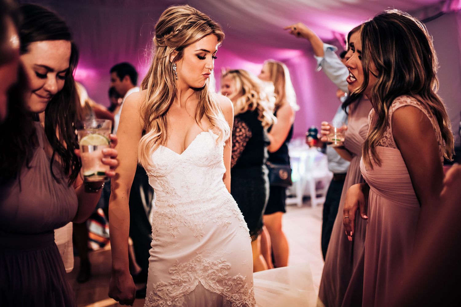 A bride in a white wedding dress dances with guests at a reception under pink lighting.
