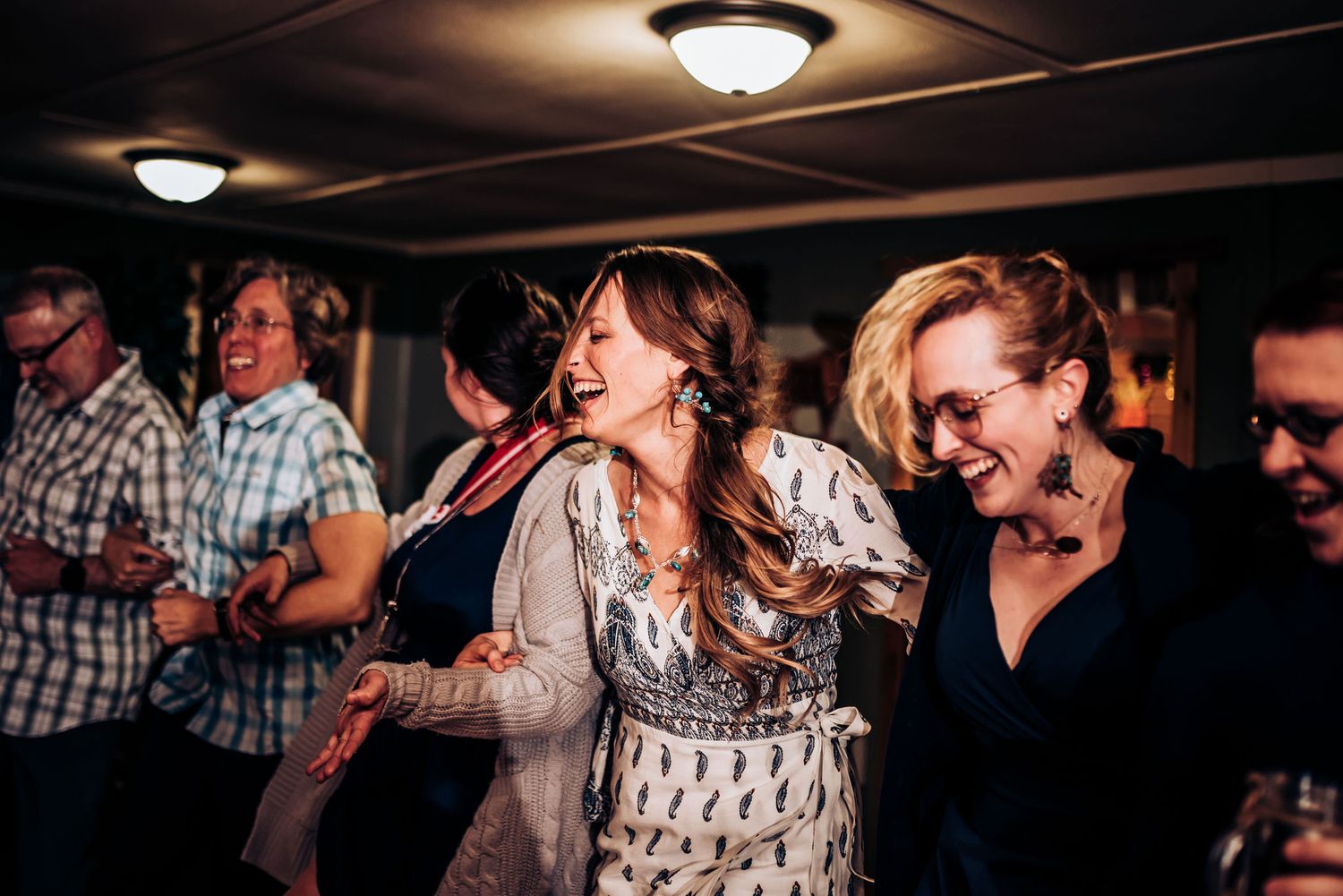 Group dances and celebrates in dimly lit room with wood paneling and ceiling lights.