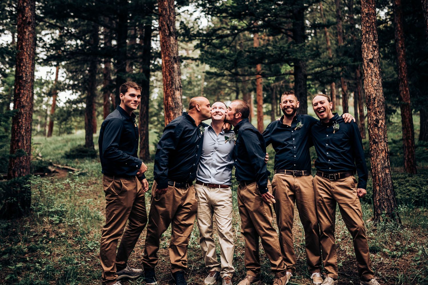 Wedding party members in coordinated outfits share lighthearted moment in pine forest.