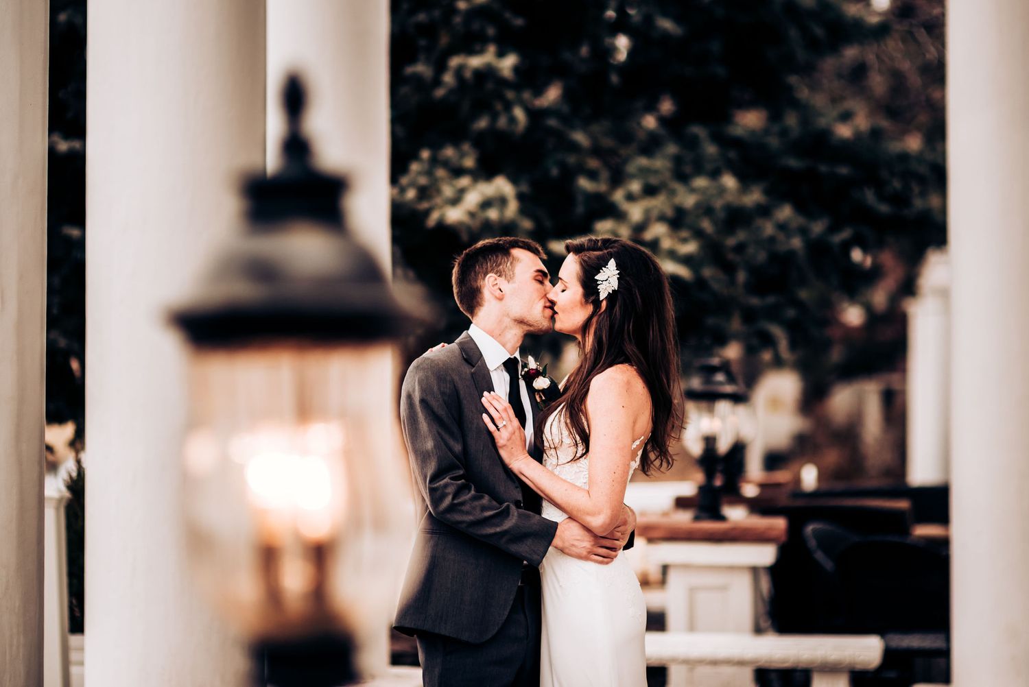 Romantic wedding kiss captured between white columns with glowing lantern in background.