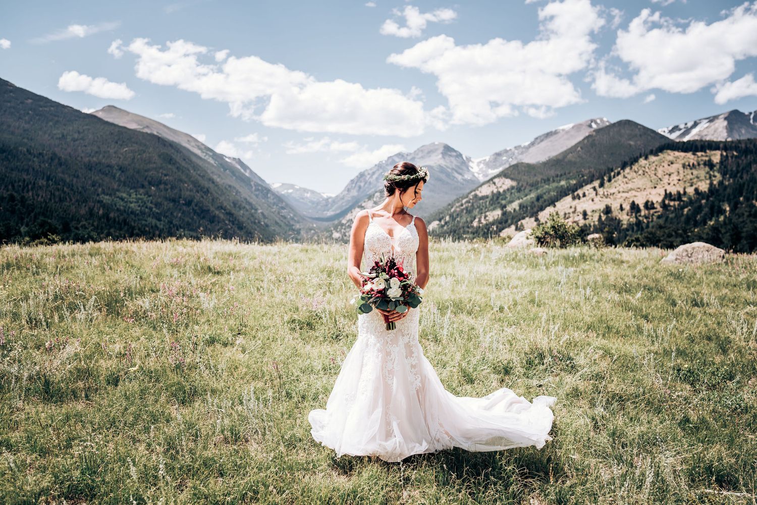 A flowing white wedding gown contrasts against majestic mountain peaks and sunlit meadow.