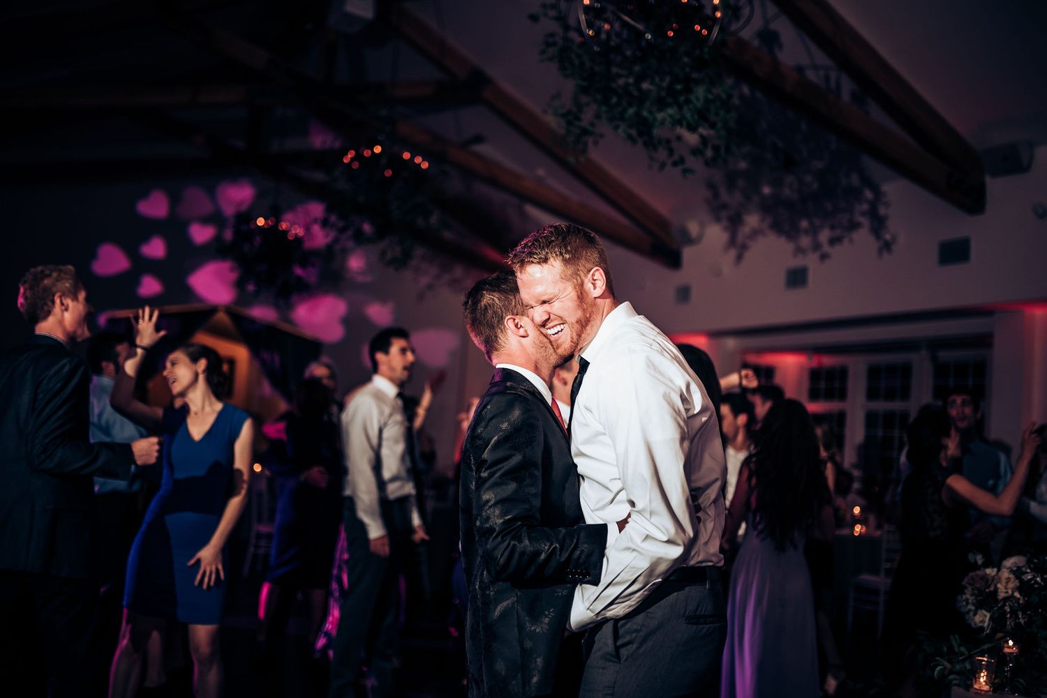 Couples slow dancing at wedding reception under romantic purple lighting and greenery.
