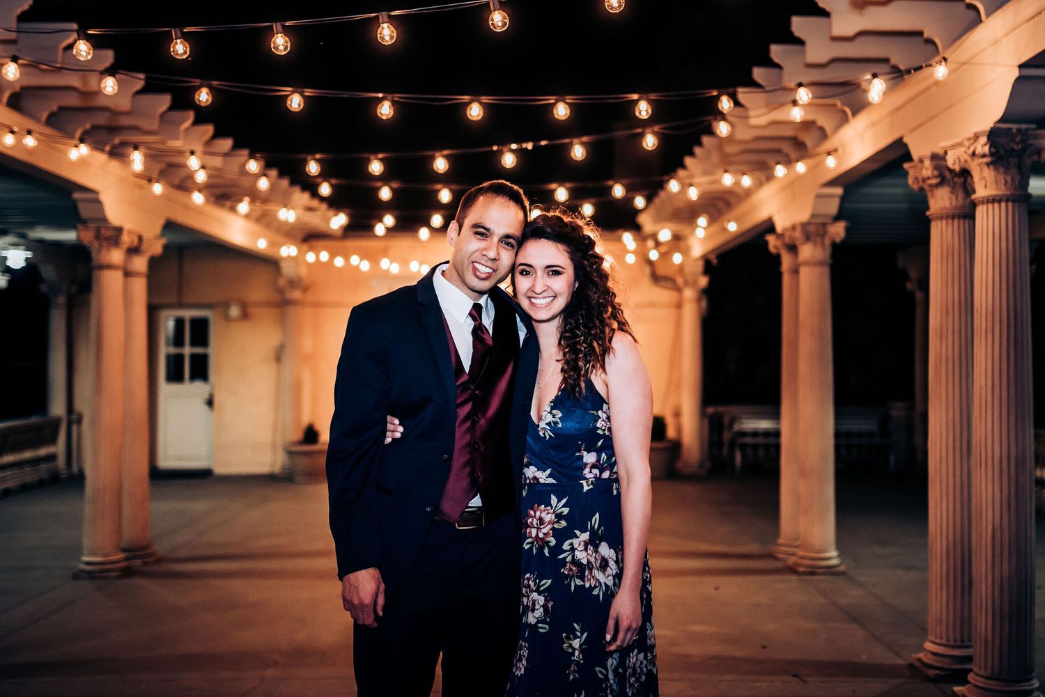 Couple poses beneath romantic string lights in an elegant columned veranda at night.