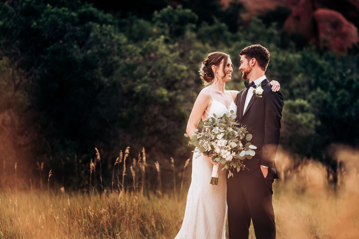 Golden sunset light illuminates wedding couple among tall grass and rocky landscape.