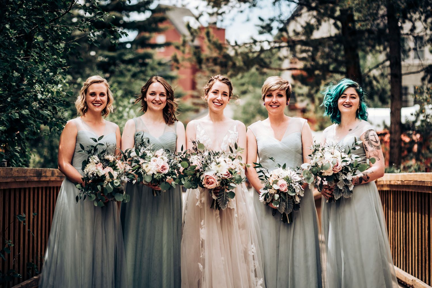 Wedding party with bridesmaids in soft gray dresses holding lush floral bouquets on wooden deck.