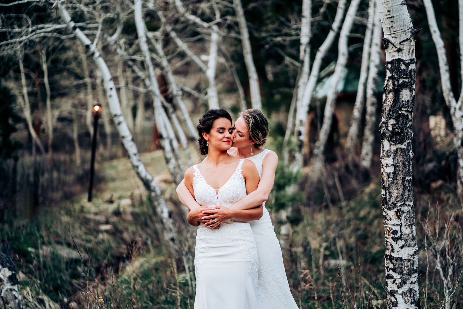 Two people in white wedding dresses embrace among birch trees in a forest setting.